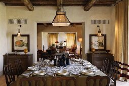 A dining room with a view into another room - a laid dining table with antique wooden chairs under a rustic wood beamed ceiling