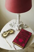 An arrangement on a white occasional table - a table lamp with a red shade, a Venetian mask and a red-bound book