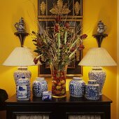 Chinesische Vase mit Plisseeschirm und Porzellan auf der Kommode mit Amaryllisstraus vor kräftigem Gelb auf der Wand