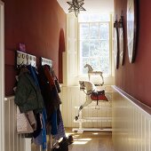 Coat hooks with children's clothes in a hallway with a dark red wall and white wooden half panelling and view onto an antique rocking horse