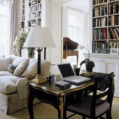 Work on a black, antique davenport with a table lamp with a white shade in front of an upholstered sofa in a library