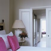 A view from a bedroom into a corridor - a table lamp with a light-coloured lamp shade next to a bed with pink cushions
