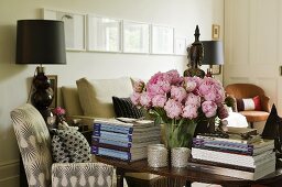 A bunch of pink peonies between two stacks of books on a table and a table lamp with a black shade next to upholstered armchair