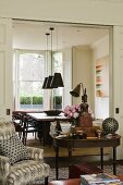 An antique wooden table and a patterned armchair in front of a doorway into a dining room with a view of a table and chairs