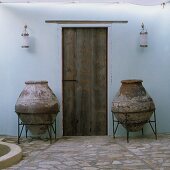 A Mediterranean courtyard - heavy amphorae in metal stands in front of a rustic wooden door on a natural stone floor