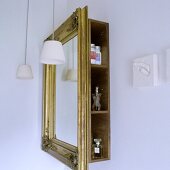 A bathroom corner - a mirror in a gold frame in front of a shelf with side opening and a pendant lamp with white glass shades