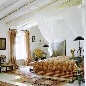 A white canopy hanging above a bed from a wood beam ceiling and antique country house-style chairs next to a window