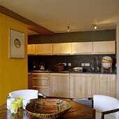 A fitted kitchen with light wooden fronts and wooden handles in front of a dark grey wall with spotlights in the ceiling