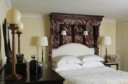 An English bedroom with table and wall lamps, a double bed with a patterned canopy and white bedclothes