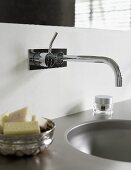 A designer wall tap over a wash basin and pieces of soap in a silver dish