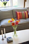 Flowers in a vase and candlesticks on a coffee table in from of a gray sofa with striped pillows