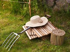 Damenhut aus Geflecht auf Holzfliesen und Mistgabel im Garten