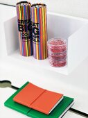 Boxes of bright colored pencils in cylindrical boxes on a white wall bracket and notebooks with coloured covers