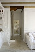 An open-plan living room with a white ladder against a cupboard and an antique Greek pillar in the hallway with an open door leading to a bedroom