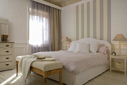 Striped wall paper at the head end of a double bed with pink bedclothes and an upholstered bench