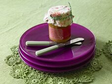 A stack of purple plate on a green tablecloth with a jar of jam and knives