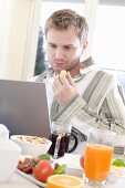 Junger Mann am Laptop beim Frühstücken
