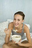 Young woman on bed, eating muesli with yoghurt