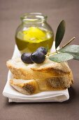 Schwarze Oliven am Zweig auf Weißbrot, dahinter Olivenöl