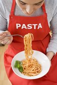 Young woman in apron eating spaghetti with tomato sauce
