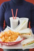 Woman holding sandwiches, cola and crisps on tray