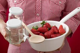 Woman holding strainer full of strawberries, jam jars, spoon