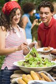 Junge Frau verteilt Blattsalat auf Teller beim Grillfest