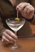 Woman holding olive on cocktail stick over glass of Martini