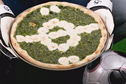 Hände halten Spinatpizza mit Mozzarella, daneben Fussball