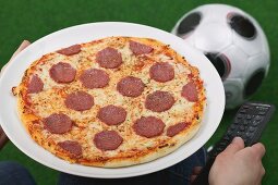 Hands holding salami pizza and remote, football