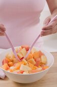 Woman taking fruit salad out of bowl with salad servers