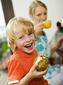 Boy holding bread roll, mother drinking coffee in background