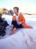 Frau sitzt im Sand, Capri-Jeans, orangeroter, knielanger Parka