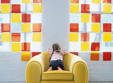 Rear view of girl sitting on armchair in front of yellow and orange window glass