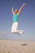 Pretty woman in blue top and white pants jumping on beach with arms outstretched, smiling