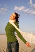 Carefree woman in green sweater, jeans and scarf enjoying sun on beach