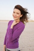 Portrait of pretty woman with windswept hair in purple sweater standing on beach, smiling