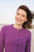Portrait of pretty woman with windswept hair in purple sweater standing on beach, smiling