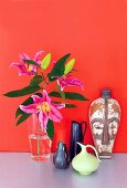 Flowers in glass vase, vases and ethnic mask on table against red wall