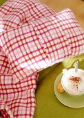 Cappuccino Tasse steht neben rot- weißem Kissen und Decke.