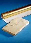 Wooden board with long nail in centre and rolled-up golden gift paper on blue background
