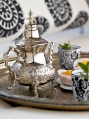 Close-up of silver tray with silver teapot, black-white glass and muffins
