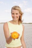 Blonde woman standing on beach and holding an orange, smiling