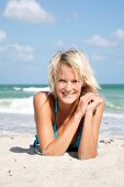 Portrait of beautiful blonde woman wearing blue bikini lying on beach, smiling