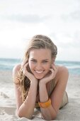 Portrait of beautiful blonde woman wearing brown top lying on beach, smiling