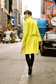 Pretty woman with short hair in yellow overcoat walking on street, looking over shoulder