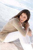 Beautiful woman wearing beige cardigan sitting on beach and looking away, laughing