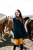 Happy woman wearing poncho and scarf standing in front of icelandic horses, laughing