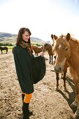 Happy brunette woman wearing poncho and scarf stroking icelandic horse and smiling