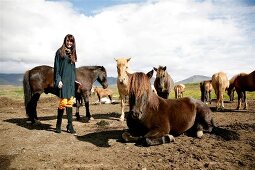 Brunette woman wearing poncho and scarf standing between many icelandic horses